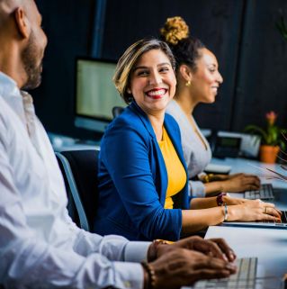 Three diverse professionals working and smiling at office desks, fostering teamwork and collaboration.